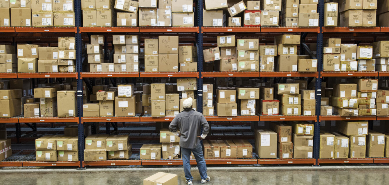 Warehouse worker checking inventory next to large racks of cardboard boxes holding product in a distribution warehouse. Warehouse worker checking inventory next to large racks of cardboard boxes holding product in a distribution warehouse.
